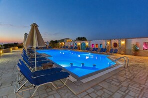 A swimming pool with lounge chairs and umbrellas, surrounded by a stone-paved deck and a building with arched windows. - Golden Bay Apartments-garden Haven (Hersonissos)