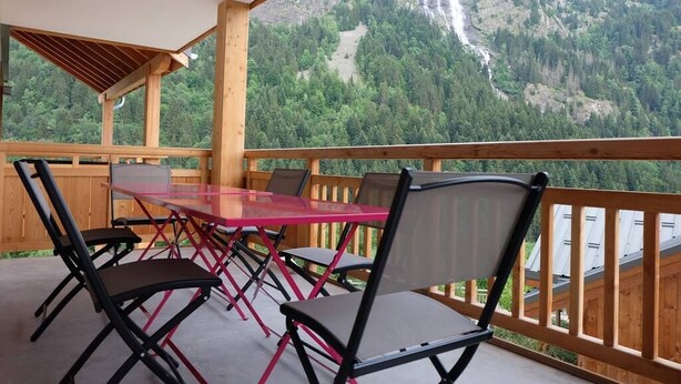 A balcony with a glass table and chairs, overlooking a mountainous landscape with a waterfall.