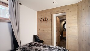 Un dormitorio con cama, una silla, paredes de madera, una puerta que da a un baño y una ventana con cortinas.