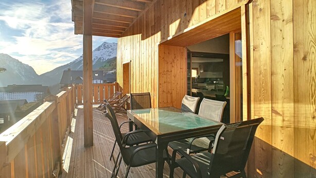 Una terraza de madera con una mesa y sillas de vidrio, con vista a un paisaje montañoso.