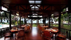 A covered outdoor dining area with wooden tables and chairs, a glass ceiling, and a view of a pool and palm trees. - Santosa Villas & Resort (Senggigi)