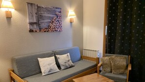 Ein gemütliches Wohnzimmer mit einem grauen Sofa, zwei Holztischen, einem Holzstuhl mit einem Fellüberwurf und einem an der Wand befestigten Kunstwerk mit aufgeschichteten Holzstämmen.