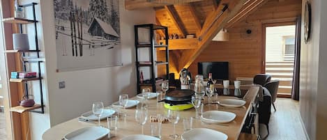 Un coin repas avec une longue table dressée pour le repas, des poutres en bois au plafond et un tableau représentant un paysage enneigé accroché au mur.