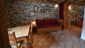 A rustic room with a stone wall, wooden ceiling beams, a wooden table, a red sofa, and a tiled floor.