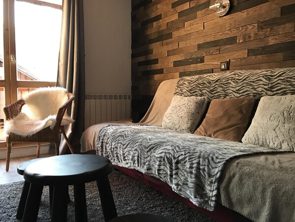 Um quarto aconchegante com parede de madeira, cama com roupa de cama estampada, uma cadeira e um banquinho pequeno.