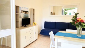 Ein Zimmer mit Fernseher auf einer Holzkommode, einem Sofa, einem Esstisch mit Stühlen und einer Vase mit Blumen.