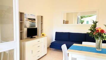 A room with a TV on a wooden cabinet, a sofa, a dining table with chairs, and a vase with flowers.