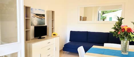 A room with a TV on a wooden cabinet, a sofa, a dining table with chairs, and a vase with flowers.