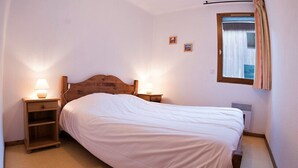 Un dormitorio con cabecera de madera, cama individual, dos mesitas de noche con lámparas, ventana con cortinas y un respiradero en la pared.