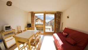Una habitació acollidora amb una taula i cadires de fusta, un sofà vermell i una porta corredissa de vidre que dona a un balcó amb vista a la muntanya.