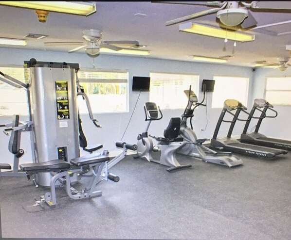 Ett gym med olika träningsmaskiner, en takfläkt och fönster med persienner.
