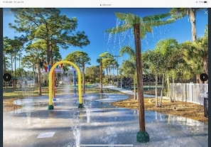 Stroll through the serene park, surrounded by lush trees and a tranquil water feature with a central archway and multiple water jets along a paved walkway.