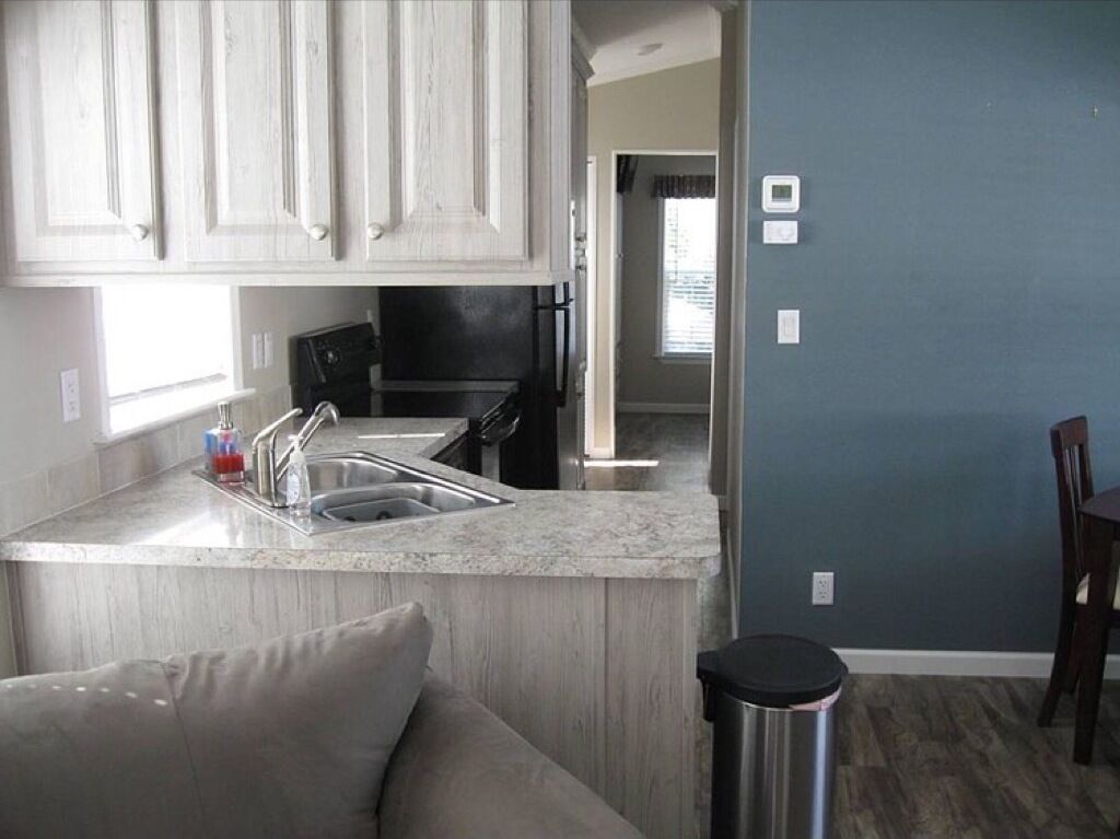 A compact kitchen with a sink, countertop, and cabinets. There is a trash can and a dining area visible.