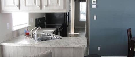 A compact kitchen with a sink, countertop, and cabinets. There is a trash can and a dining area visible.