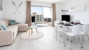 En moderne stue med sofa, spisebord med stole, fjernsyn og udsigt over byen gennem store vinduer.