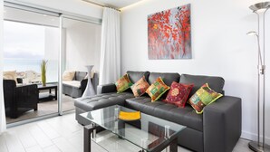 Un salon moderne avec un canapé en cuir noir, des coussins colorés, une table basse en verre et un grand tableau abstrait accroché au mur.