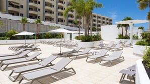 Un espace piscine aménagé sur le toit, avec des chaises longues et des parasols, entouré de palmiers et d’un immeuble à plusieurs étages.