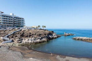 A coastal hotel with a rocky shoreline, clear blue sea, and a paved walkway leading to the beach.