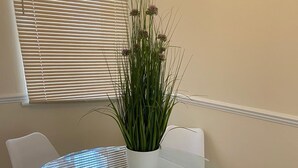 A round glass table with four white chairs, a potted plant, and a window with blinds. - Reading 3 Bedroom apartment (Reading)