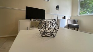 A modern living room with a white table, a black geometric sculpture, a flat-screen TV on a cabinet, a floor lamp, and a window with a view of greenery. - Reading 3 Bedroom apartment (Reading)