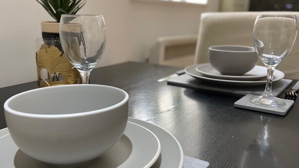 A dining table set with white plates, bowls, and cutlery, with a plant and decorative items in the background. - Modern 1 Bed Apartment (Reading)