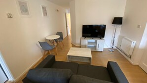A modern living room with a black sofa, a white coffee table, a TV on a white stand, and a dining area with a small table and chairs. - 3 Bedroom Apartment (Reading)