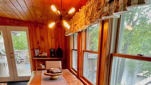 Ambiente interno in legno con tavolo da pranzo, sedie e vista su un lago attraverso ampie finestre.