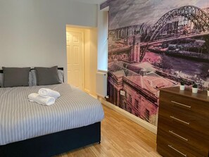 Une chambre à coucher avec un lit, des oreillers, un radiateur, une commode en bois et une fresque représentant un paysage urbain sur le mur.