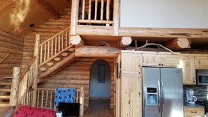 Interior de una cabaña de madera con una escalera que lleva a un balcón, una zona de cocina con nevera y microondas, y una mesa con mantel azul.