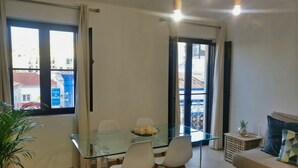 Un salón moderno con una mesa de comedor de cristal, sillas blancas, un sofá con cojín y una planta en maceta.