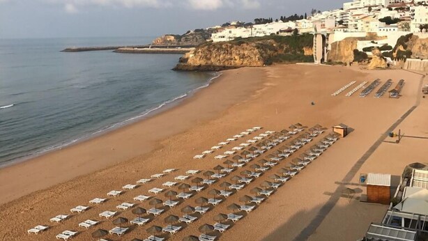Una playa con filas de tumbonas y sombrillas, un litoral rocoso y edificios al fondo.