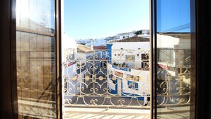 Vista a través de una ventana con celosías de metal ornamentales, un edificio con fachadas azules y blancas, y un cielo despejado.