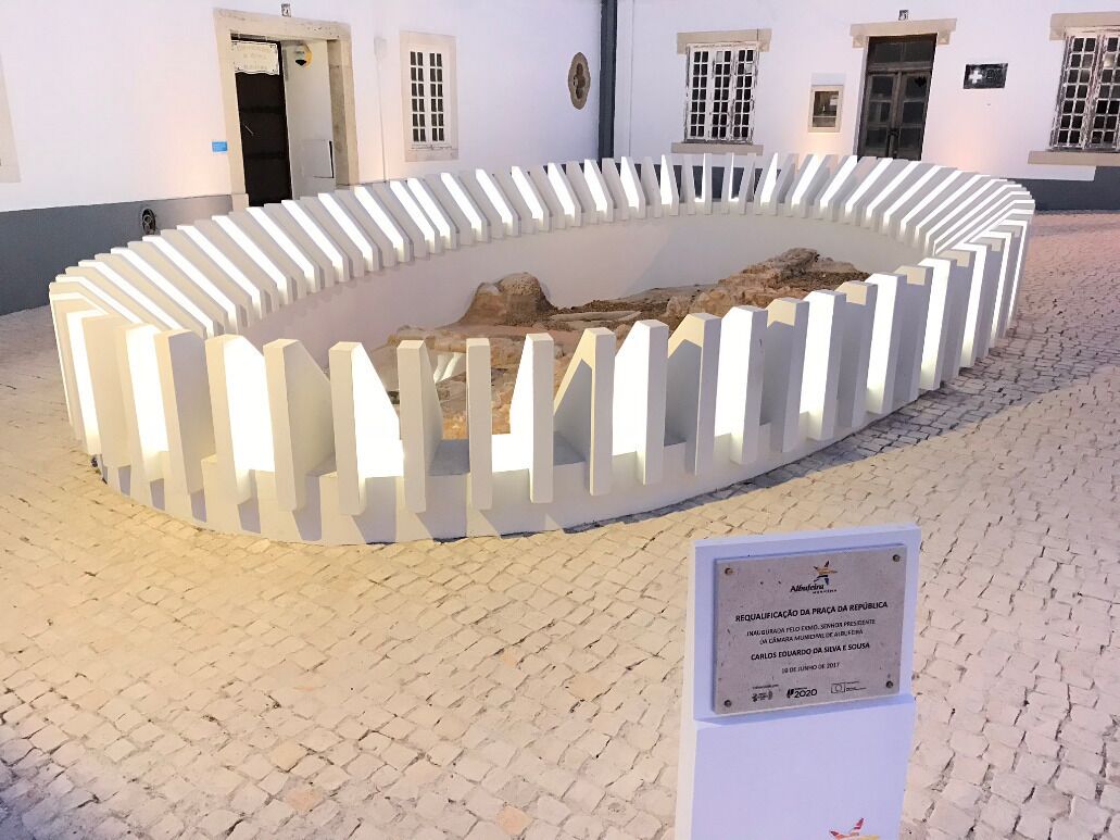 Une exposition circulaire, avec une structure en forme de clôture blanche, une plaque commémorative et un sol pavé de pierres.