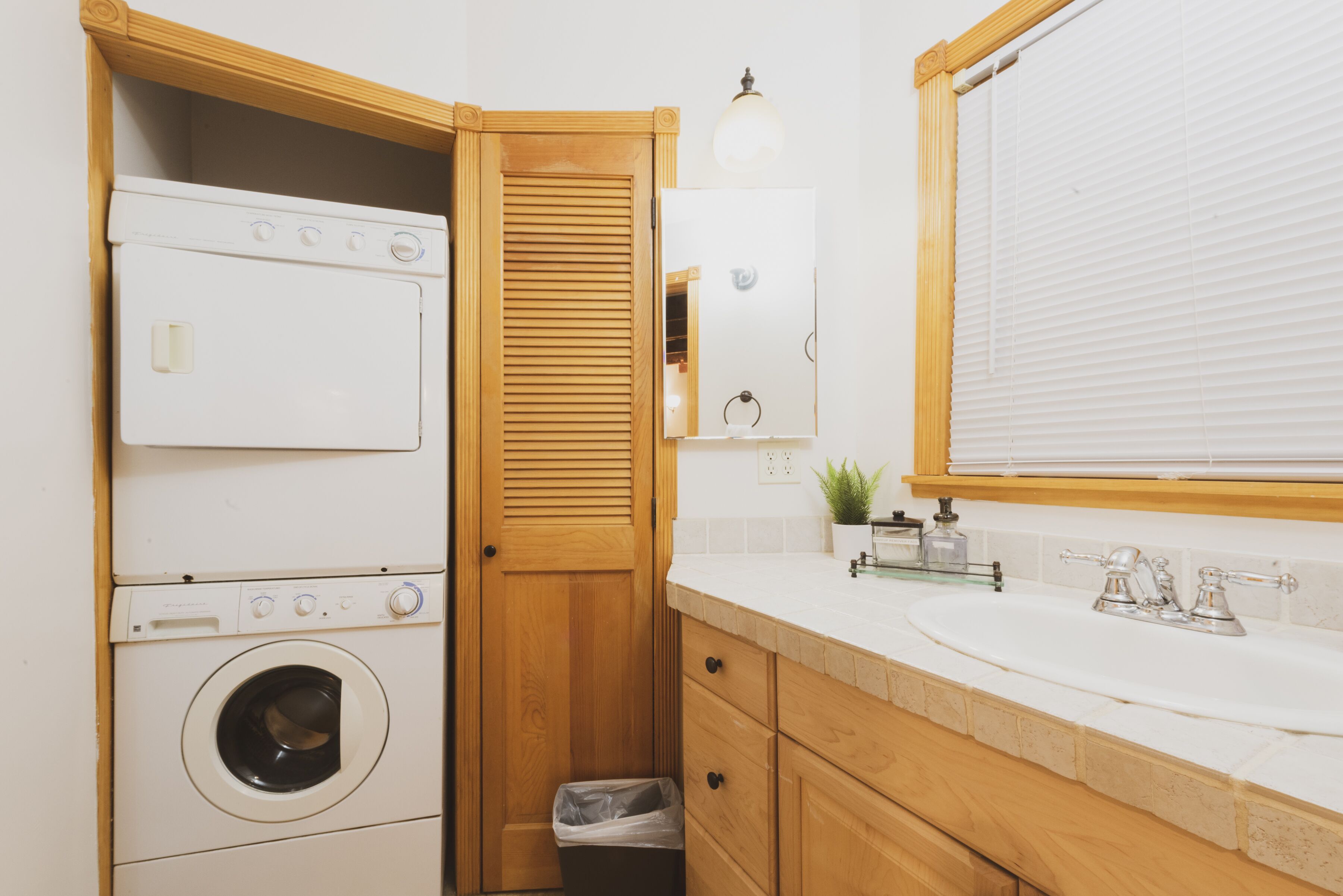 Een kleine wasruimte met een wasmachine en droger, houten kastjes, een kleine prullenbak, een spiegel en een wastafel met kraan.