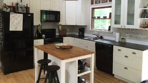 A modern kitchen with white cabinets, a black refrigerator, a wooden island with stools, and a window with greenery outside. - Cozy Cabin Getaway - Convenient to 4 Ski Areas, Hiking and Mountain Bike Trails (Davis)