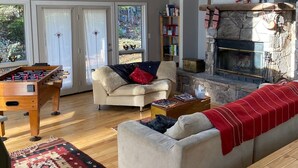 A cozy living room with a foosball table, a fireplace, and a large window with curtains. - Cozy Cabin Getaway - Convenient to 4 Ski Areas, Hiking and Mountain Bike Trails (Davis)