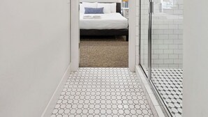 Un pasillo estrecho con una cama al fondo, un ventilador de techo y una zona de ducha con azulejos blancos.