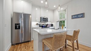 Una cocina moderna con armarios blancos, electrodomésticos de acero inoxidable, una isla blanca con dos sillas de mimbre y un letrero que dice 'vida en el lago'.