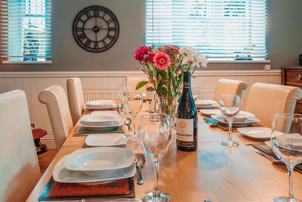 Una mesa de comedor dispuesta con platos, vasos y una botella de vino, con un reloj y flores en la pared.