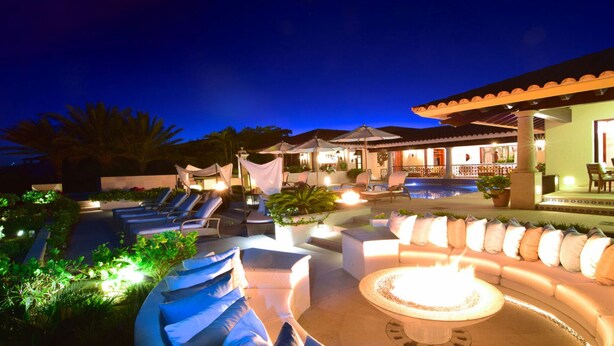 Un espacio exterior bien iluminado con sillones, una fogata y una piscina.