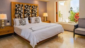 Una habitación de hotel con una cama grande, dos mesitas de noche con lámparas, un cabecero de madera con piedras decorativas y vistas a un balcón con plantas.