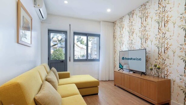 A modern living room with a yellow sofa, a wooden TV stand, and a wall with a bamboo wallpaper pattern. - Anna´s Flats | Jardim do Morro by HostWise (Vila Nova de Gaia)
