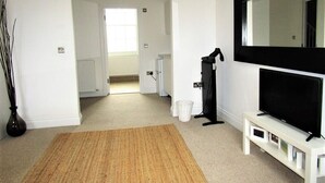 A living room with a large TV, a white bench, a black stand, and a woven rug. - Large Private Double En-suite - 5 (London)