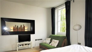 A bedroom with a bed, a green armchair, a television on a stand, a window with curtains, and a lamp. - Large Private Double En-suite - 5 (London)