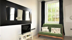 A room with a green sofa, a flat-screen TV on a white stand, a large window with curtains, and a mirror on the wall. - Large Private Double En-suite - 5 (London)