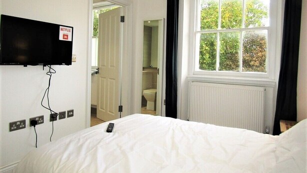 Habitación de hotel con cama, televisor montado en la pared, ventana con cortinas y vista a zonas verdes.