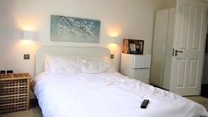 Una cama bien hecha con sábanas blancas, un control remoto sobre la cama, una mesita de madera y una lámpara adosada a la pared, además de una puerta a la derecha.