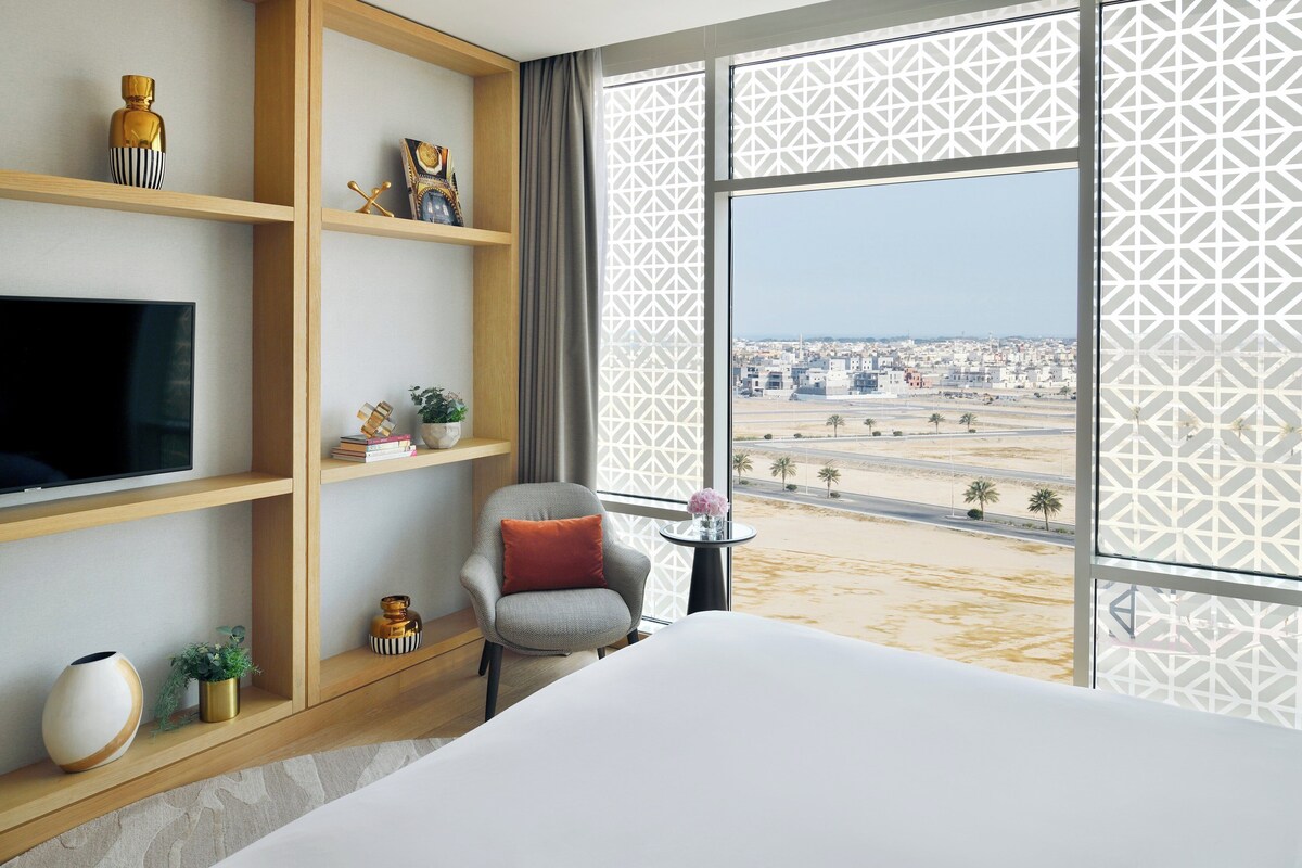 Habitación de hotel con una cama grande, televisión, una silla con cojín, una mesita con un florero y vistas a un paisaje urbano a través de cortinas estampadas.