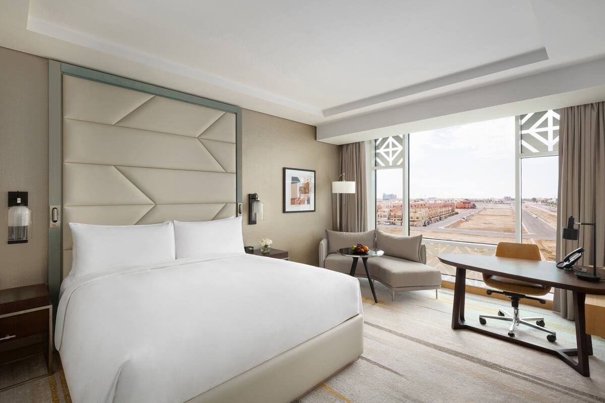 Une chambre d’hôtel avec un grand lit, un bureau, une chaise et une vue sur la ville grâce à de grandes fenêtres.