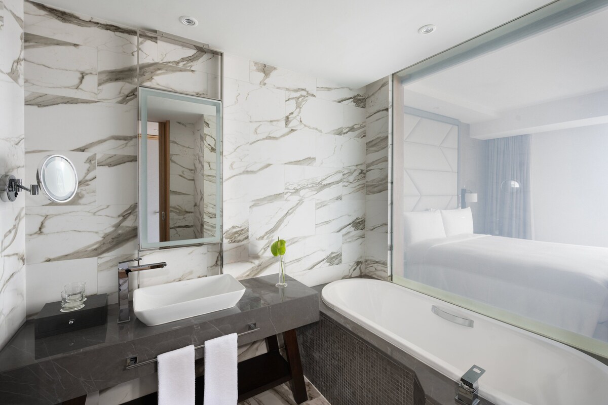 Un baño moderno con salpicadero de mármol, un espejo grande, un lavamanos, una bañera y vistas de la habitación del hotel.